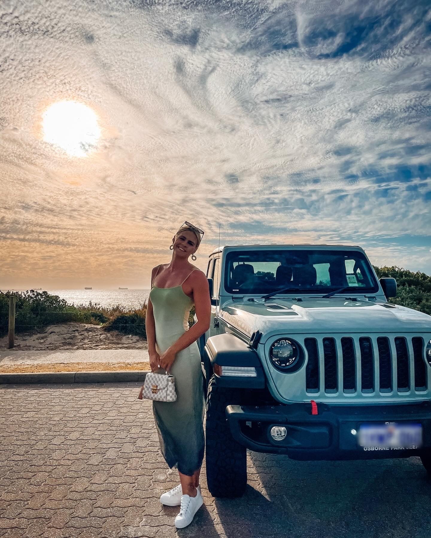 Shayna Jack standing beside a Jeep Wrangler on a coastal road at sunset.