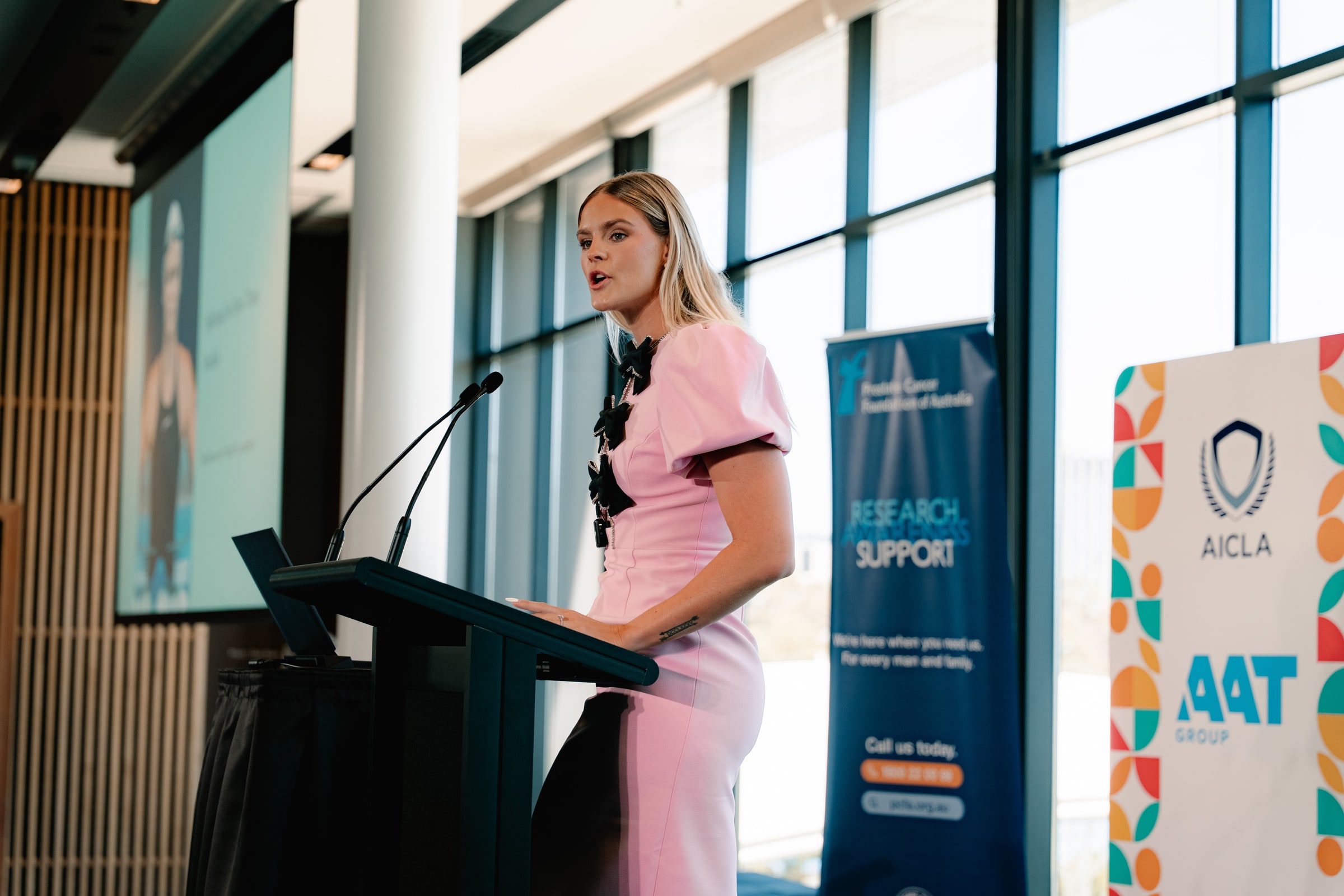 Shayna Jack mid-sentence at a lectern, with conference banners and a presentation screen behind her.