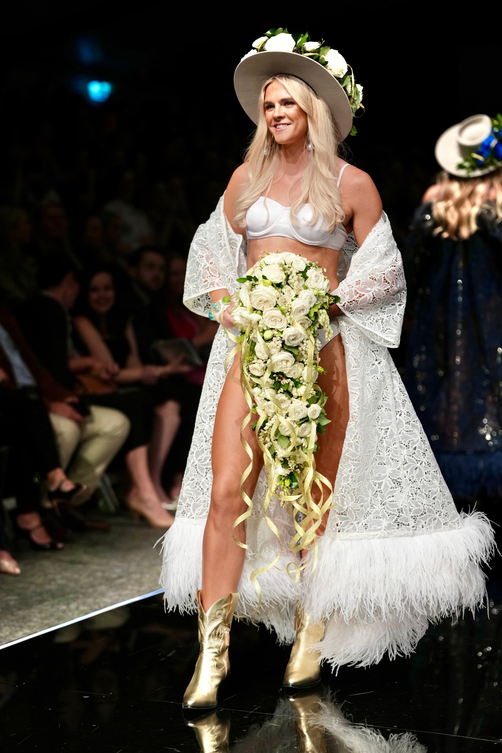 Shayna Jack walking a runway in a white outfit and wide-brimmed hat, carrying a floral bouquet.